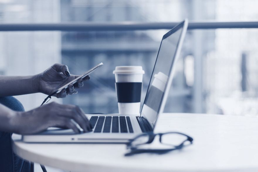Une main tape sur le clavier d’un ordinateur portable, l’autre tient un téléphone, à côté d’une paire de lunettes posée sur la table.