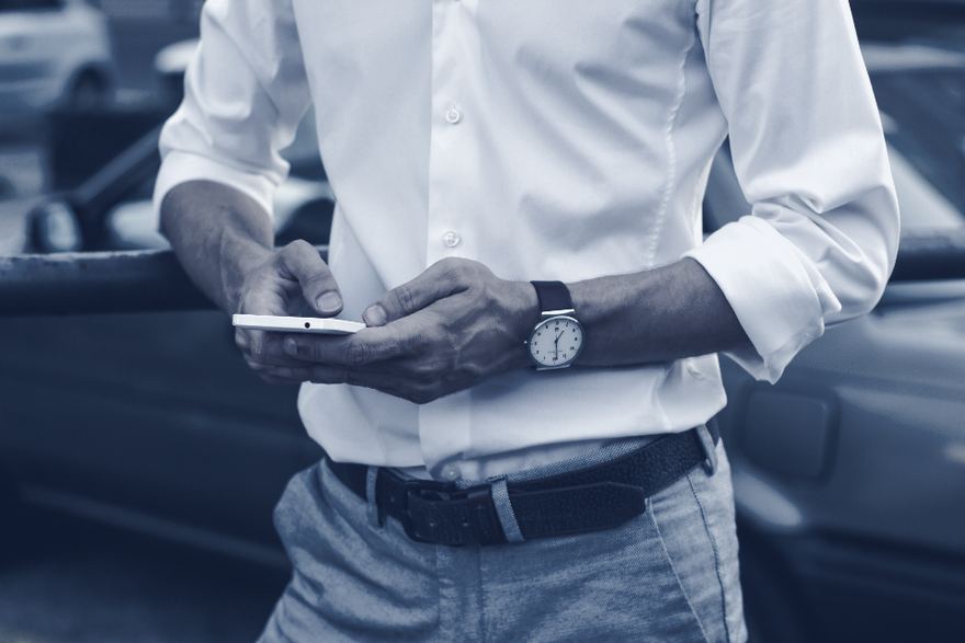 Un homme debout, vu du buste, tient son téléphone à deux mains et s’arrête avant de toucher l’écran.